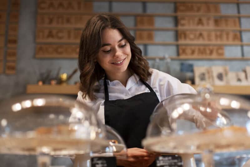 Young woman smiling behind bakery display with cakes in glass covers.