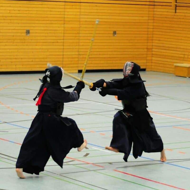 Two kendo practitioners sparring in a gym with wooden walls.