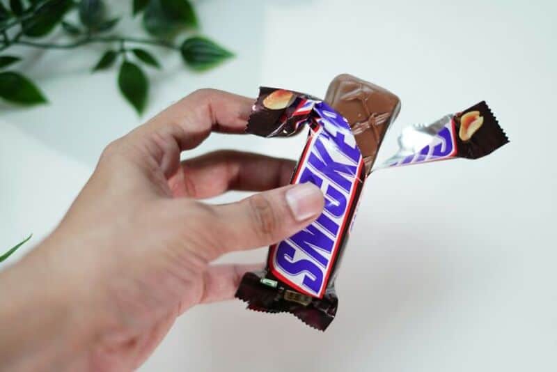 Hand holding a Snickers chocolate bar with a piece broken off.