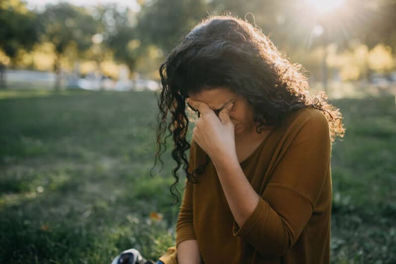 Woman sitting outside with hand on face, appearing upset, in a park at sunset.