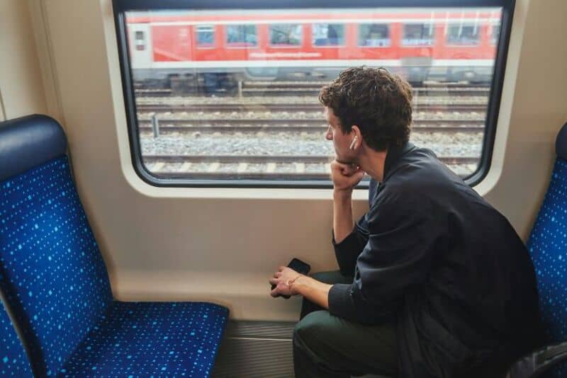 Man sitting on train, looking out window at passing train tracks and red train.