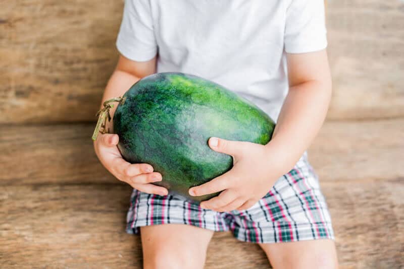 Child holding a large green watermelon sitting on a wooden surface.