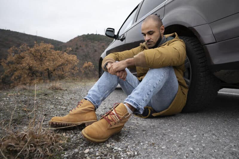 Man sitting on the ground next to a car, looking down, in a rural area.