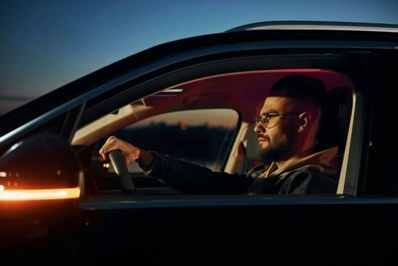 Man driving a car at sunset with a focused expression.