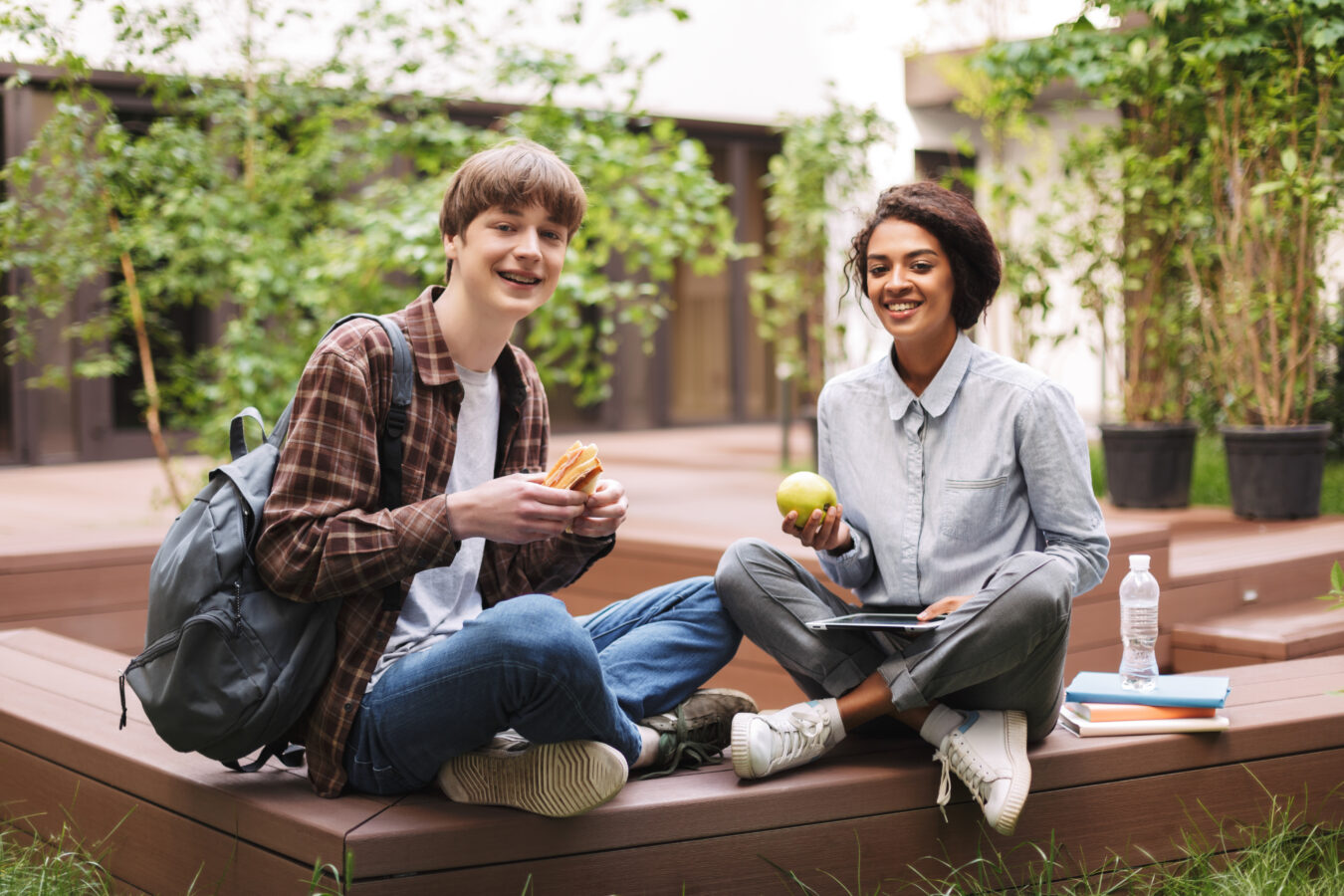 Two smiling students sitting on a wooden platform outdoors, holding a sandwich and an apple.