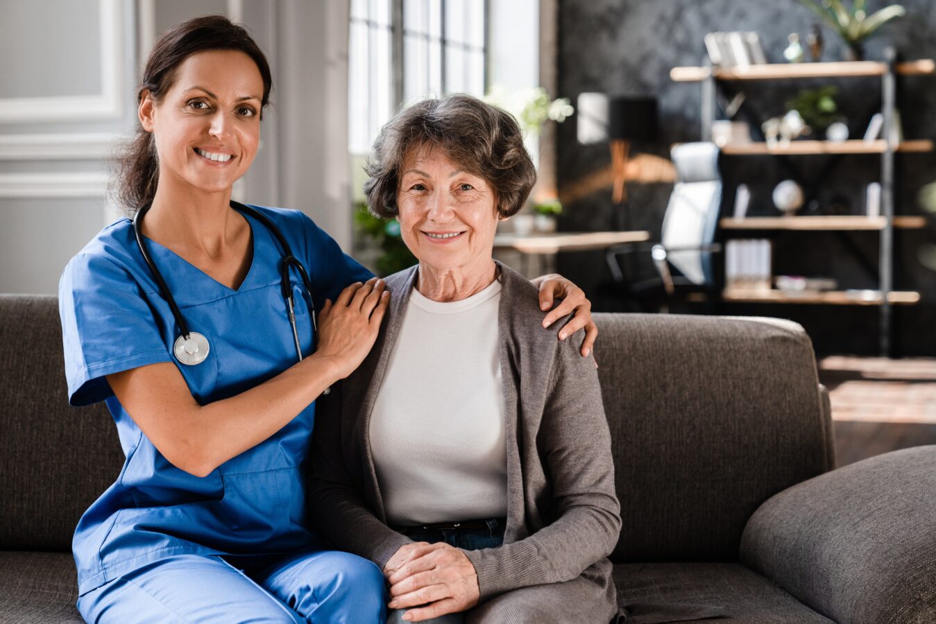 Nurse with stethoscope smiling with elderly woman on sofa.