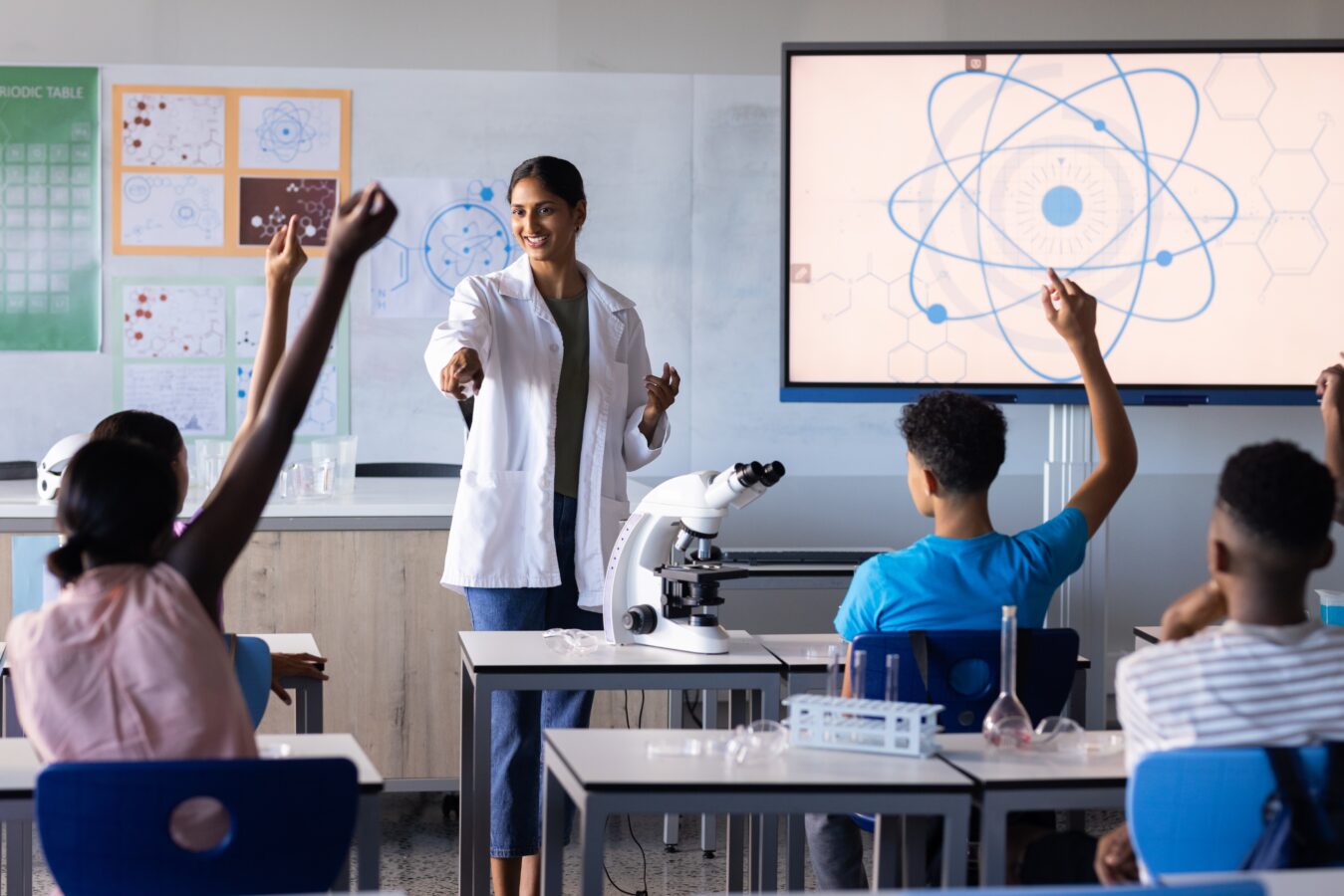Teacher smiling in science classroom with students raising hands.