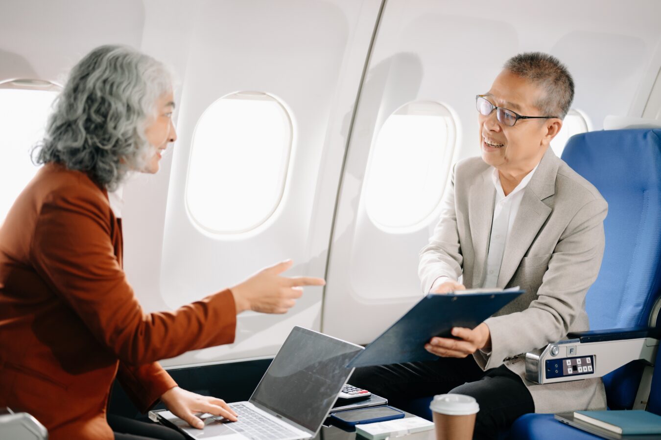 Two men having a conversation on an airplane, one with a laptop and the other with a clipboard.