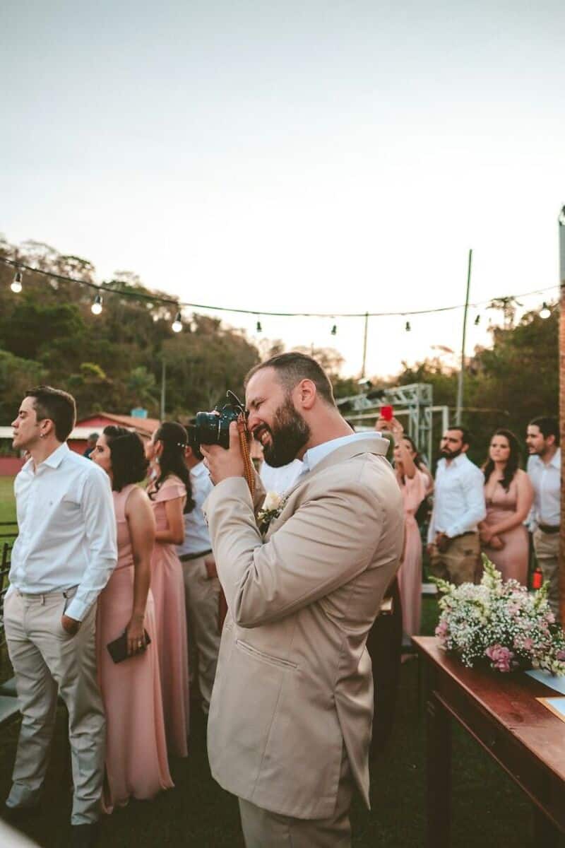 Man with beard in beige suit taking photo at outdoor wedding reception.