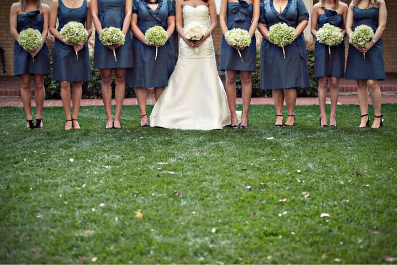 Bride and bridesmaids standing on grass, holding bouquets, at a wedding.