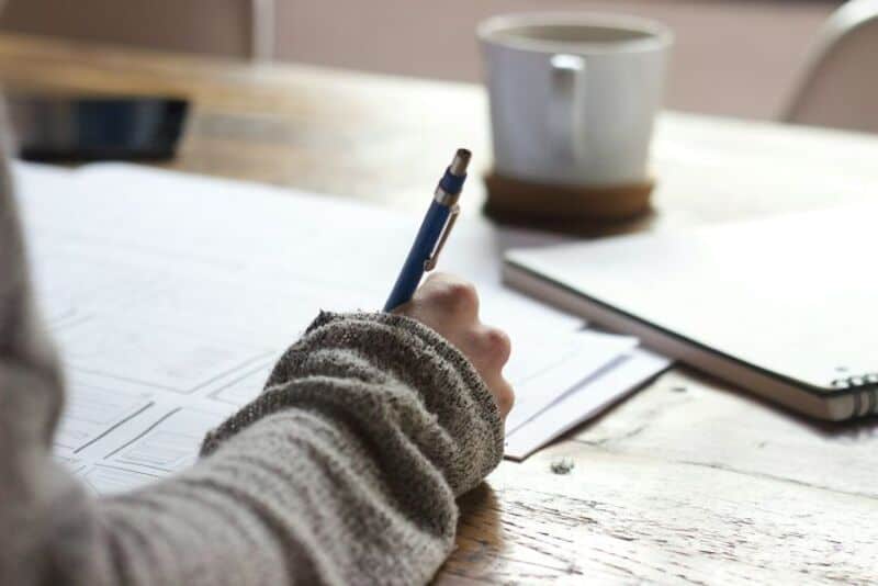 Person writing on paper with a pen, a mug and a notebook on the table.