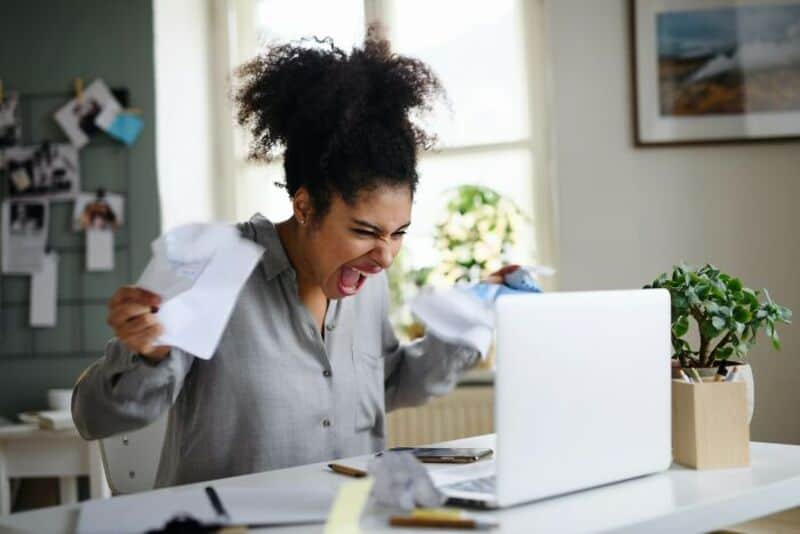 Woman frustrated at desk with crumpled papers and laptop.