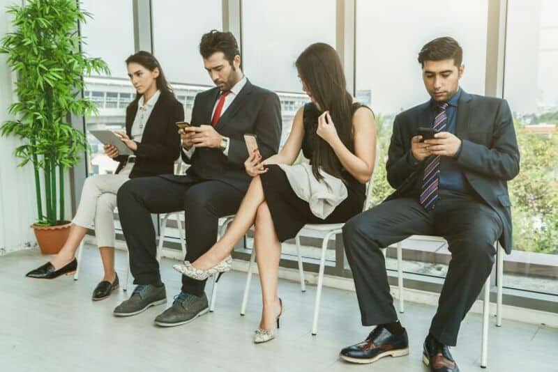 Four people sitting on chairs, all looking at their phones in a modern office.