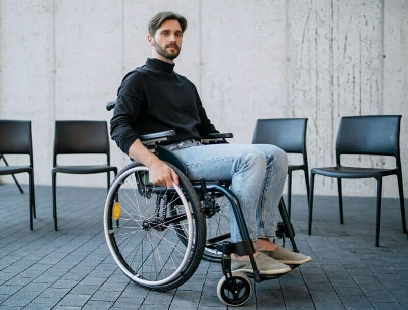 Man in wheelchair sitting outdoors with chairs behind him.