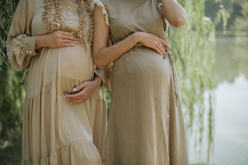Two pregnant women in beige dresses standing outdoors by a lake.