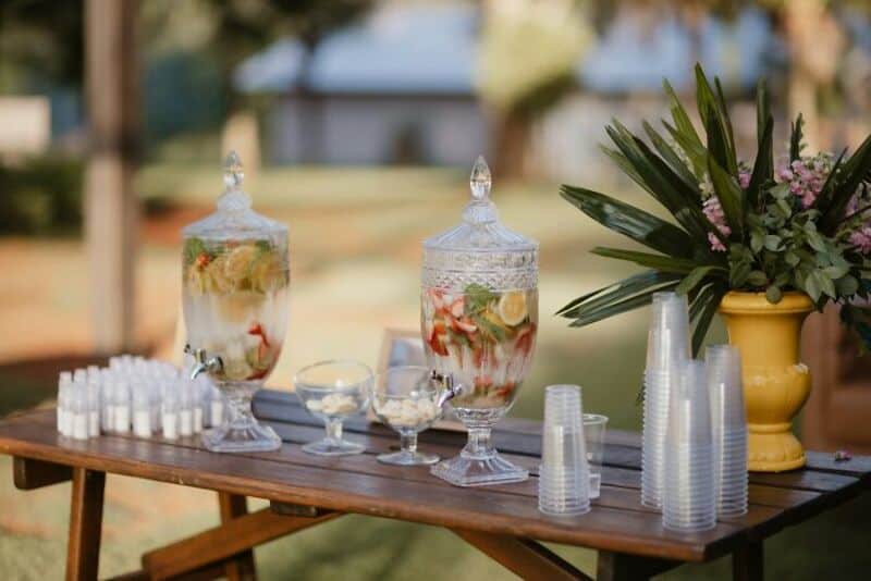 Water dispensers with fruit slices and plastic cups on a wooden table at an outdoor event.
