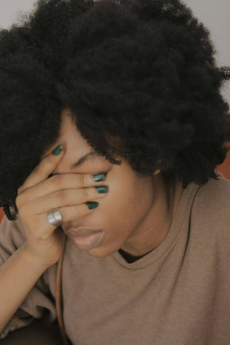 Woman with curly hair covering her face with her hand, wearing a beige top.