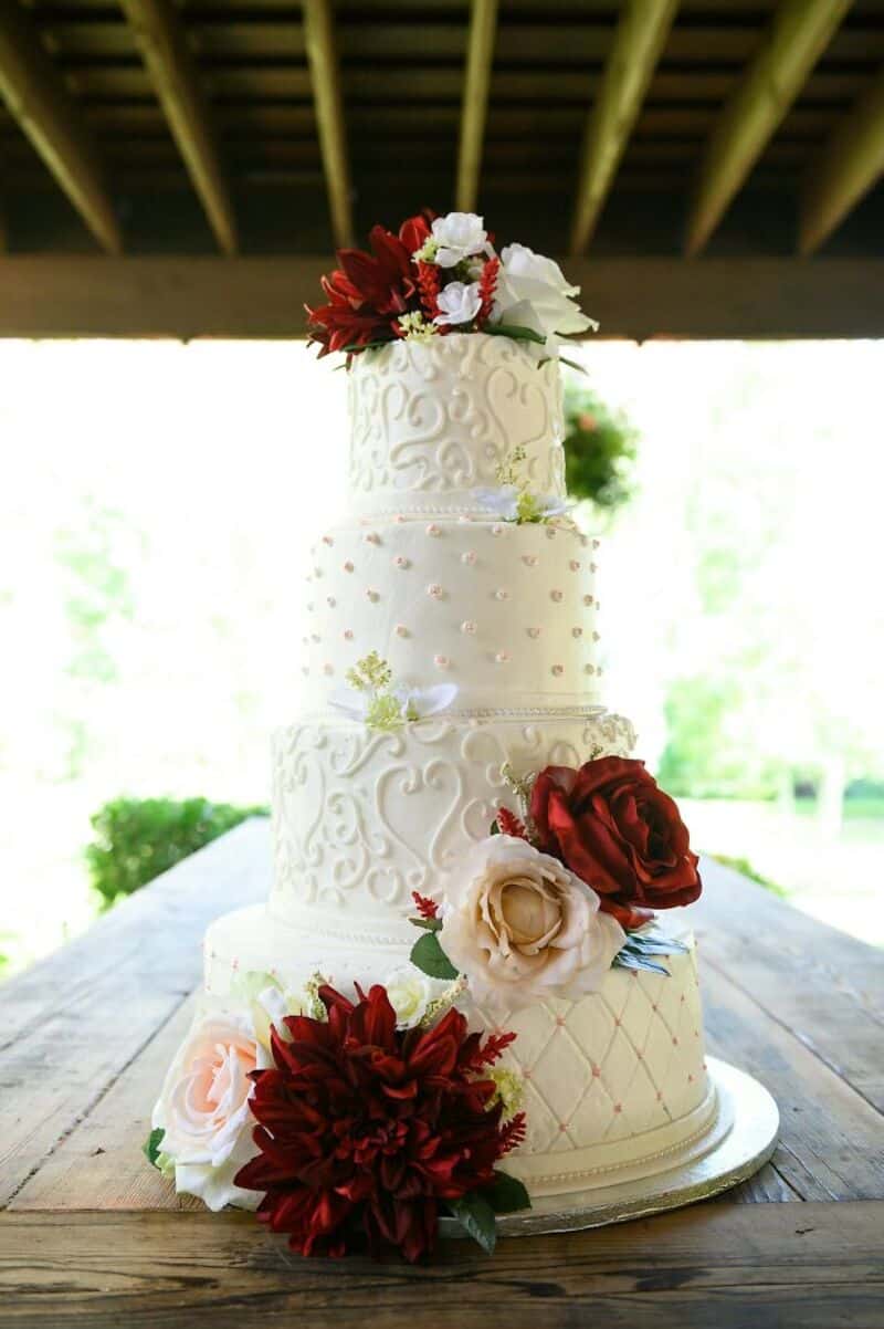 Elegant white wedding cake with red, white, and blush flowers, under a wooden pavilion.