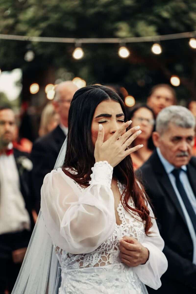 Bride crying at her wedding, surrounded by guests, outdoor setting with lights.