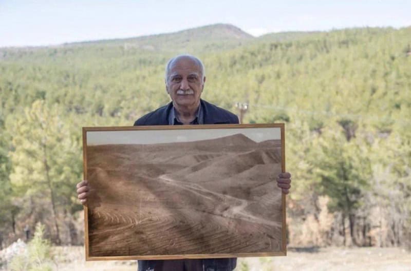 Older man holding a framed photo of a desert landscape outdoors.