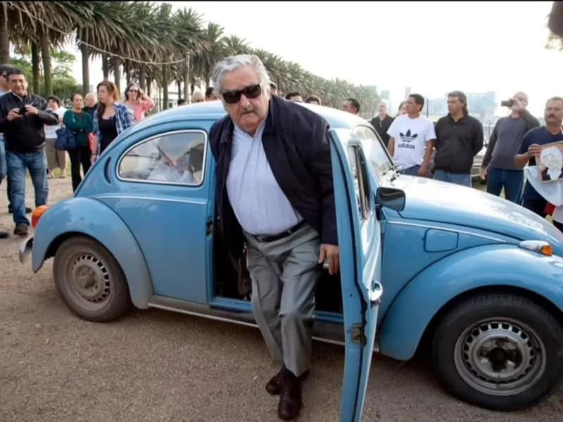 Older man with sunglasses getting out of a blue vintage car, crowd in background.