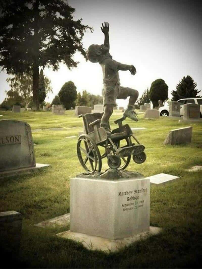 Statue of a girl in a wheelchair with one arm raised, in a cemetery.
