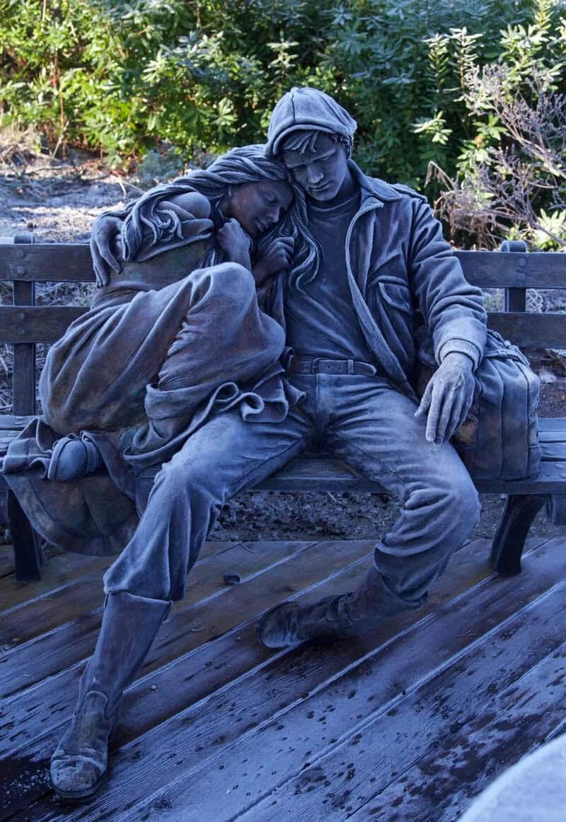 Statue of two people sitting on a bench, one resting head on the other's shoulder.