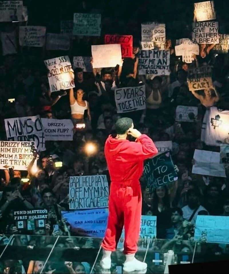 Crowd protest with signs, person in red hoodie on stage, holding microphone.