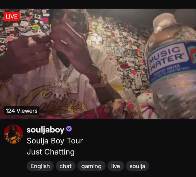 Soulja Boy in a room with a wall covered in stickers, drinking water, live stream.