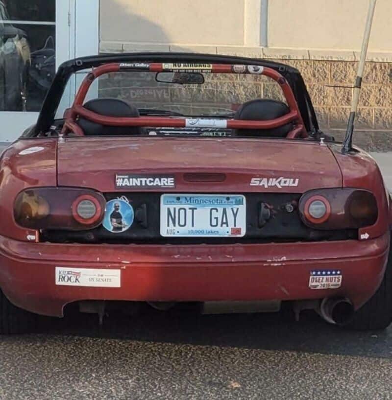 Red convertible with a Minnesota license plate reading 'NOT GAY' and various stickers.