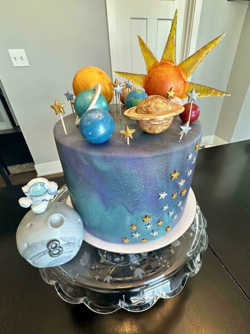 Space-themed cake with planets, stars, and a sun, on a black table.