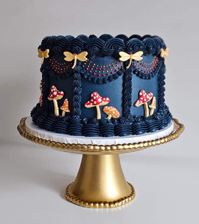 Decorative cake with blue frosting, mushrooms, butterflies, and red dots on a gold stand.