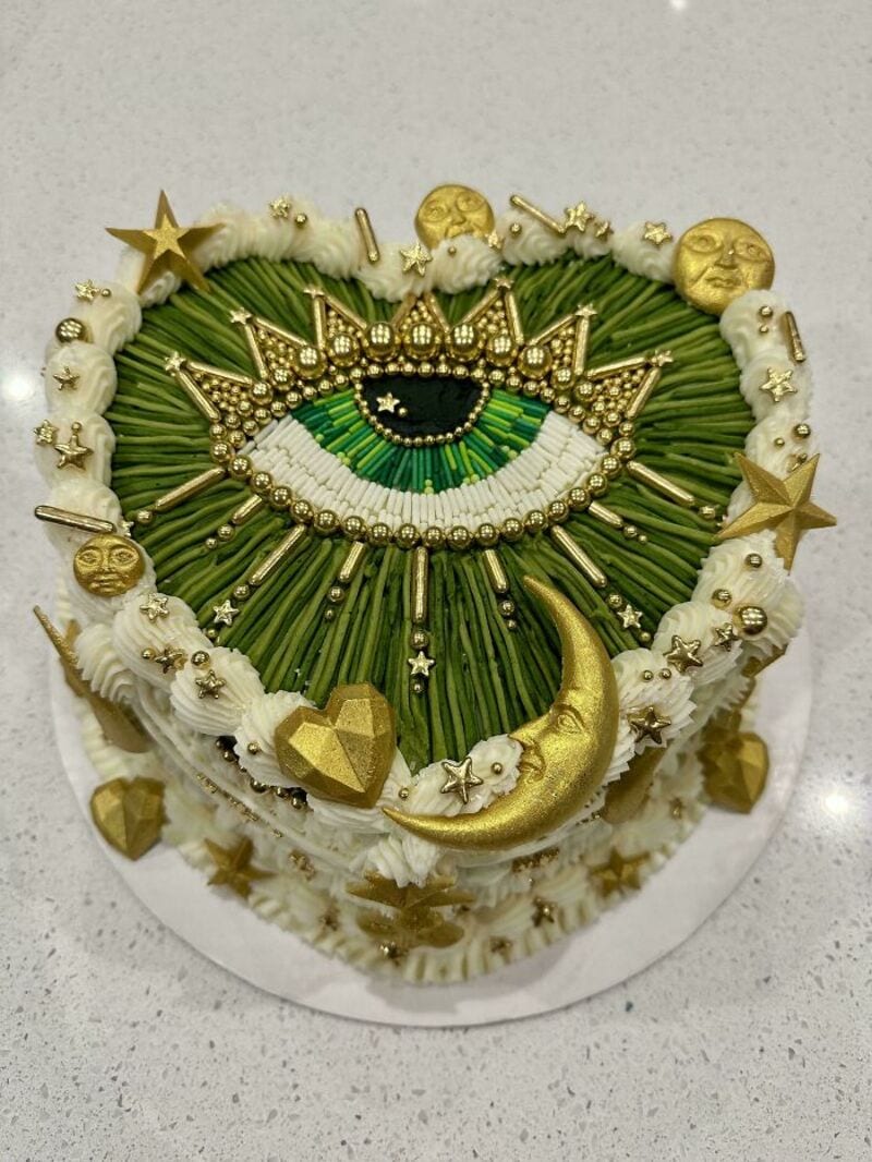 Decorative cake with an eye, stars, moon, and celestial motifs in gold, green, and white.