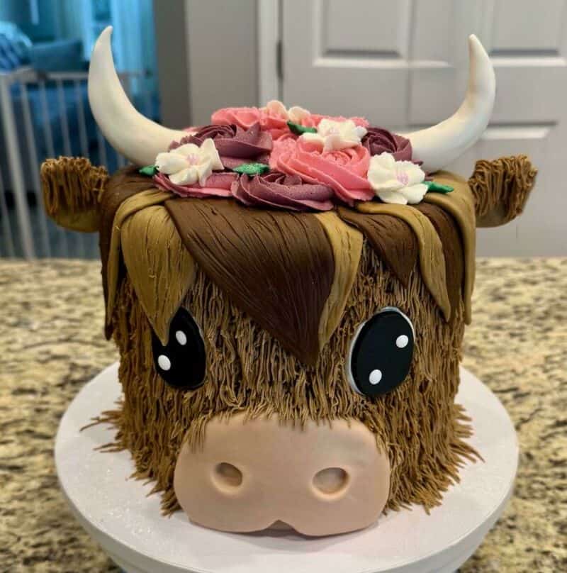Cake shaped like a bison with horns, flowers, and big eyes, on a white cake board.
