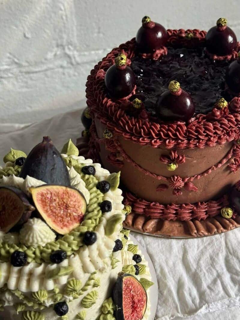 Two decorated cakes, one with cherries and gold accents, the other with figs and berries.