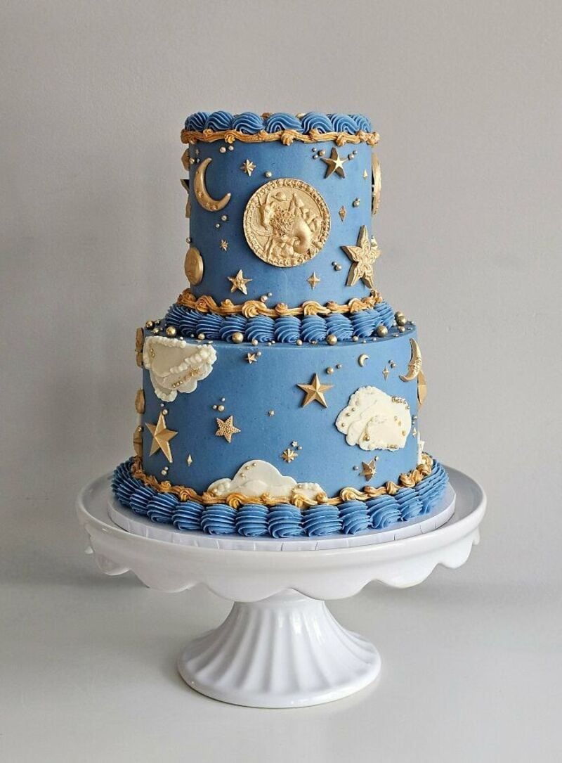 Two-tiered blue cake with gold moon, stars, clouds, and coin decorations on a white cake stand.