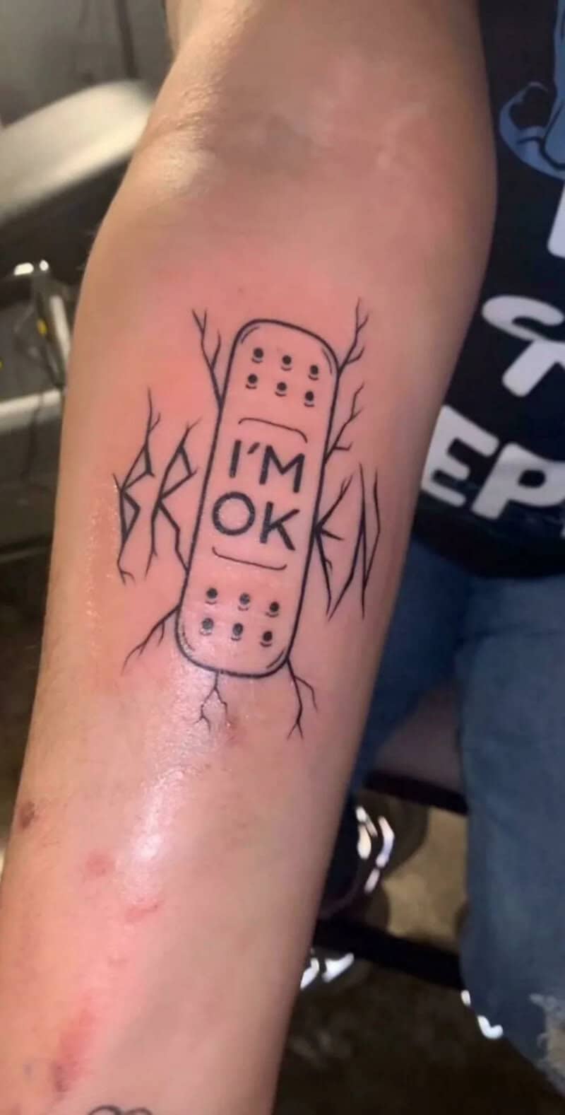 Tattoo of a bandage with 'I'm OK' on arm, surrounded by cracks.
