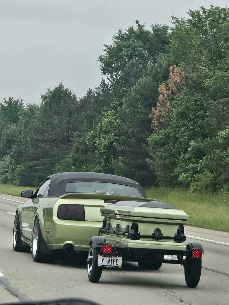 Green convertible car towing a small boat with a custom license plate 'X WIFE' on a highway.