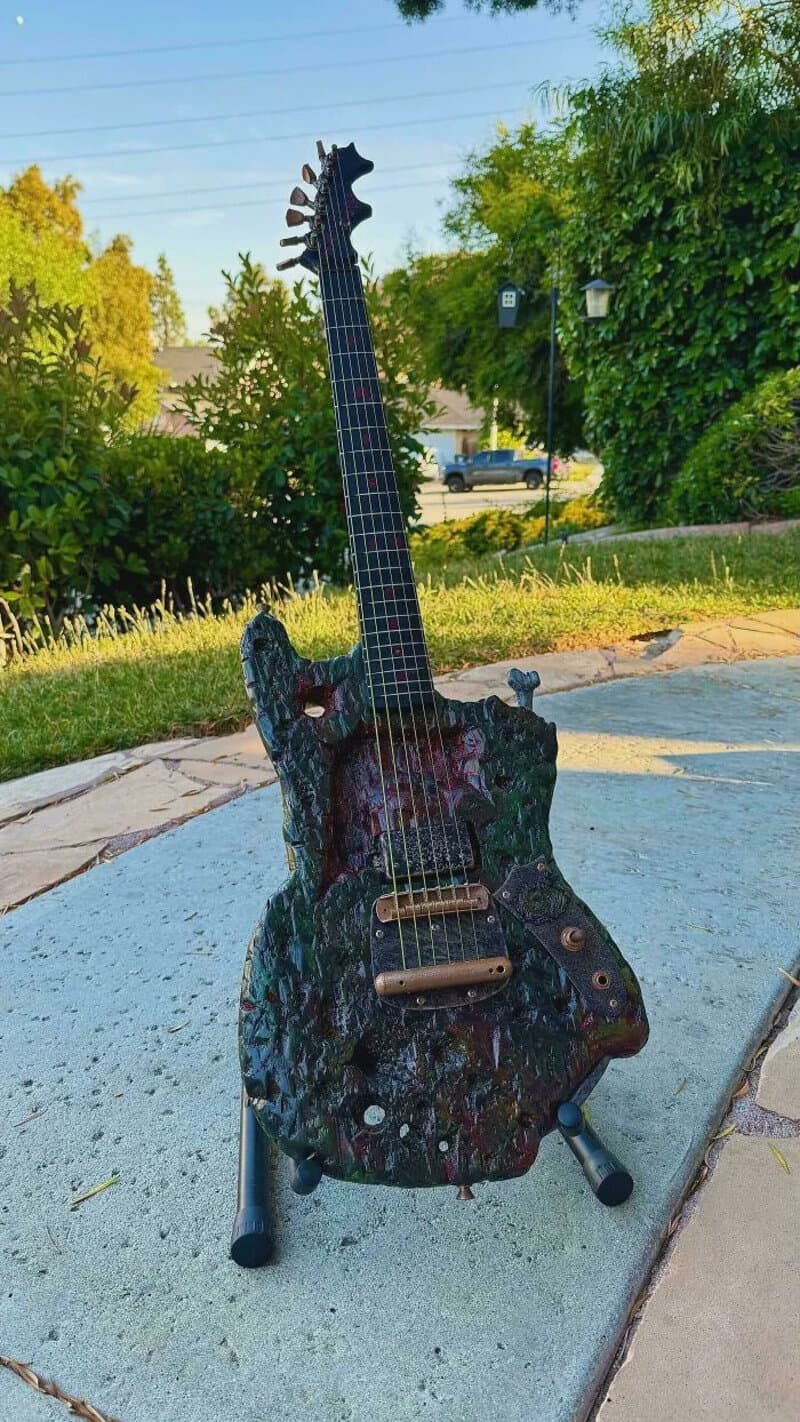 Guitar-shaped sculpture with a camouflage pattern, standing outdoors on a sidewalk.