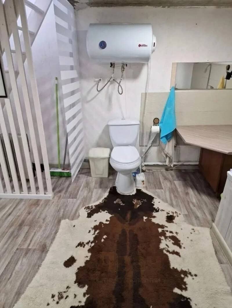 Toilet with a water heater above, brown stain on the floor rug, and a blue towel hanging.