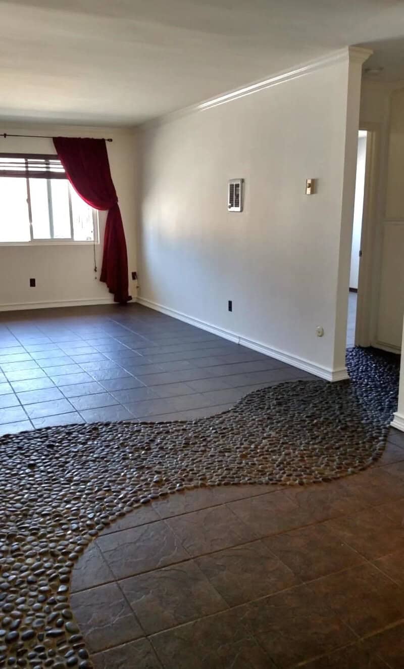 Room with a pebble pathway dividing tiled and carpeted areas, window with red curtain.