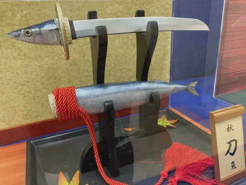 Fish mounted on a sword with a knife and red rope, display case with autumn leaf and sign.