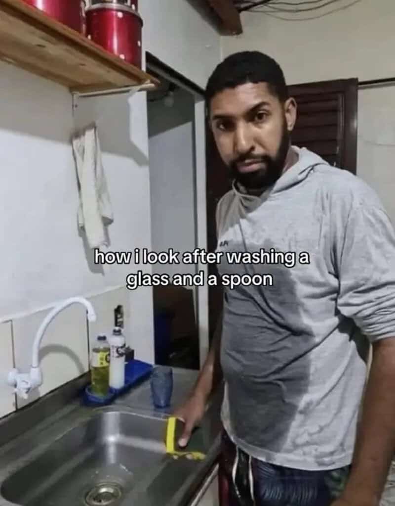 Man with a surprised expression washing a glass and a spoon at the sink.