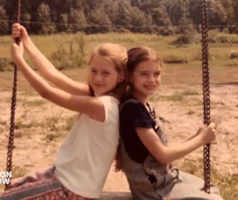 Two young girls sitting on a swing, back to back, smiling outdoors.