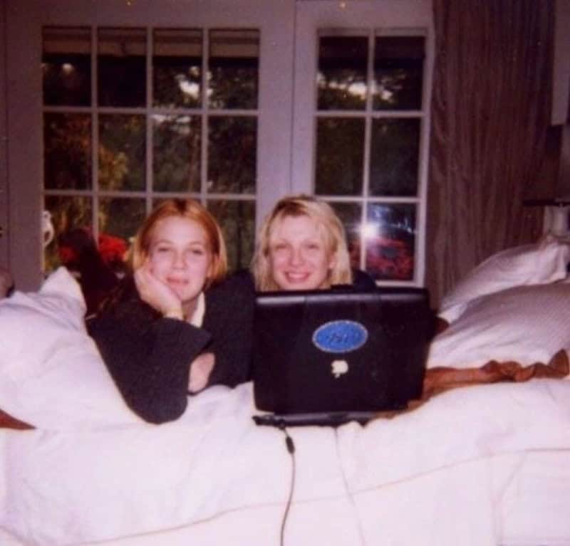 Two young girls with blonde hair using a laptop on a bed, in front of a window.