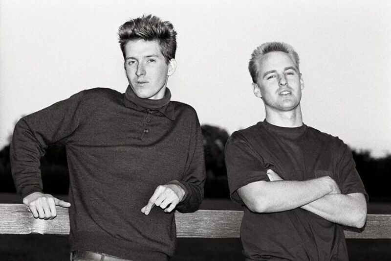 Two young men leaning on a railing, one pointing at the other, in black and white.