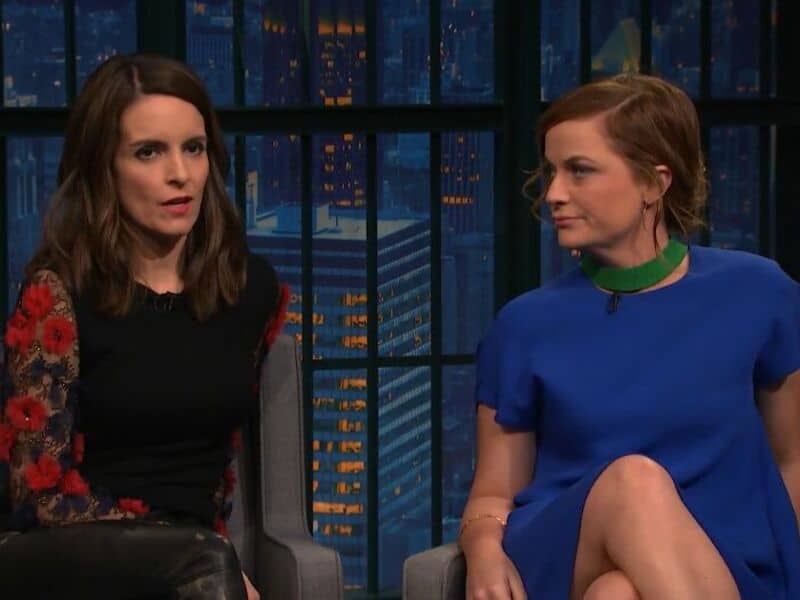 Two women sitting on a talk show set, one in a black and red dress, the other in a blue dress.