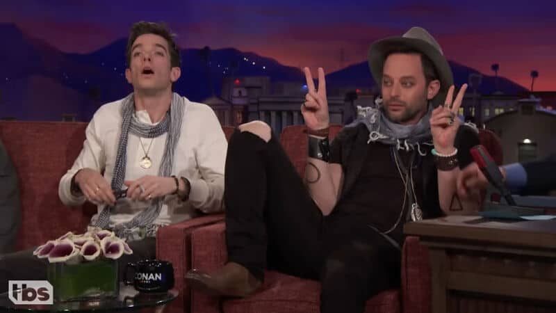Two men sitting on a talk show set, one making a peace sign, the other with a surprised expression.