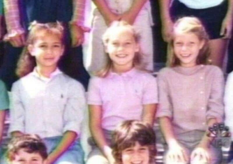 Three young girls smiling, sitting in a group photo, with other children in the background.