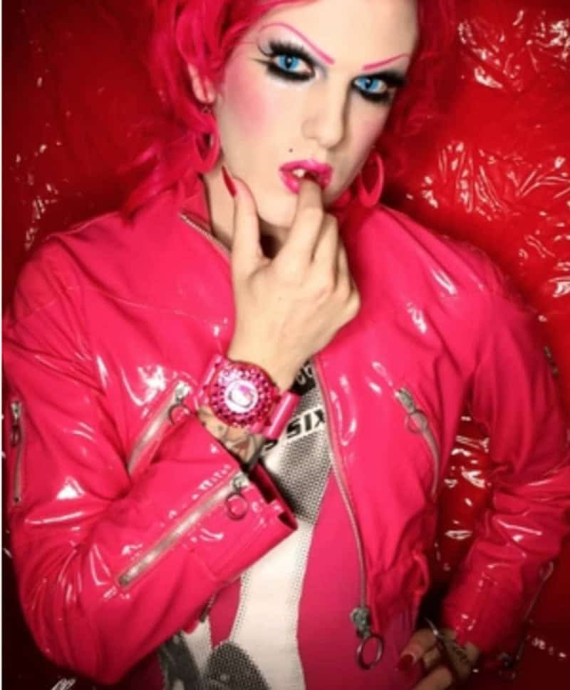 Woman with bright pink hair and makeup in a pink jacket, posing with finger near lips.